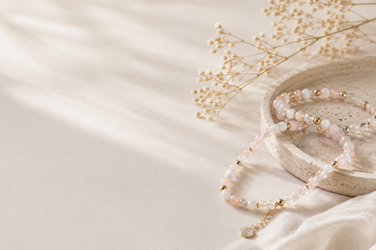 A minimalist flat lay featuring soft pastel beaded bracelets with gold accents placed on a ceramic tray over neutral fabric, styled with warm natural lighting and delicate dried flowers.